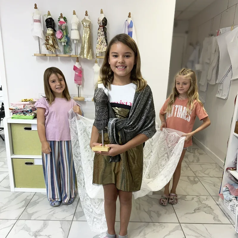 Three happy girls proudly displaying their handmade fabrics and dressed mannequins after a kids fashion sewing camp session